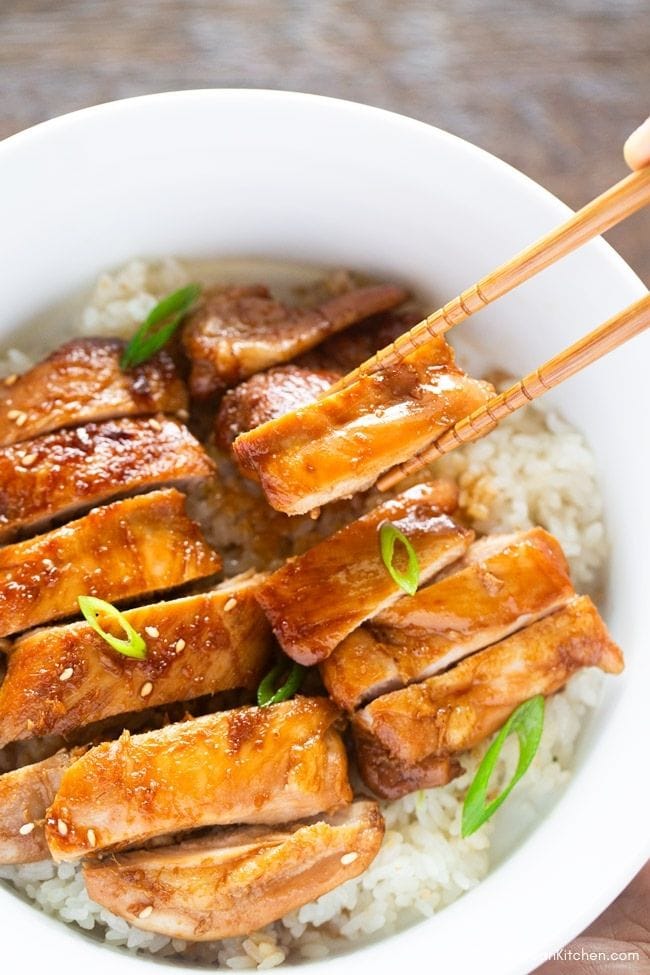 Holding a slice of teriyaki chicken with chopsticks over a plate of arranged teriyaki chicken.