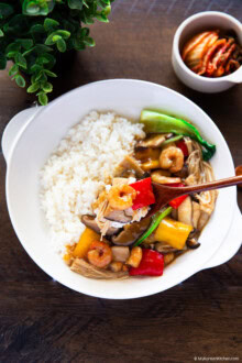 Holding a bite-sized portion of shrimp, red bell pepper, mushrooms, and rice on a wooden spoon.