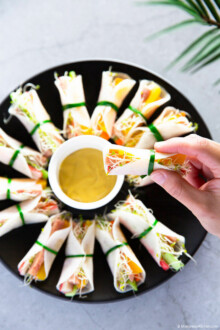 Holding a pickled radish wrap in one hand, in the background, radish wraps are served on a black plate.
