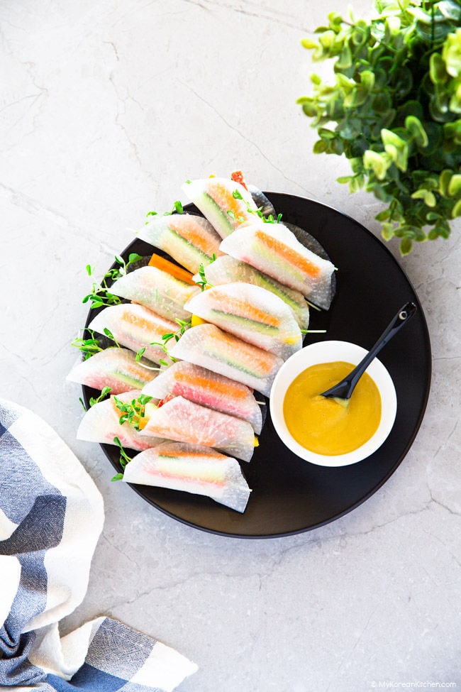 Radish wraps served on a black plate with a dipping sauce. In the background, there is a green plant.