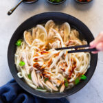 Kalguksu noodles are being held with chopsticks over a black bowl of soup, onto which red sauce is being sprinkled.