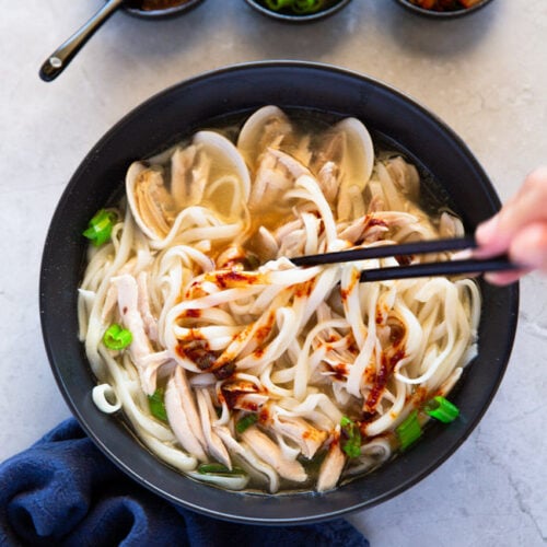 Kalguksu noodles are being held with chopsticks over a black bowl of soup, onto which red sauce is being sprinkled.