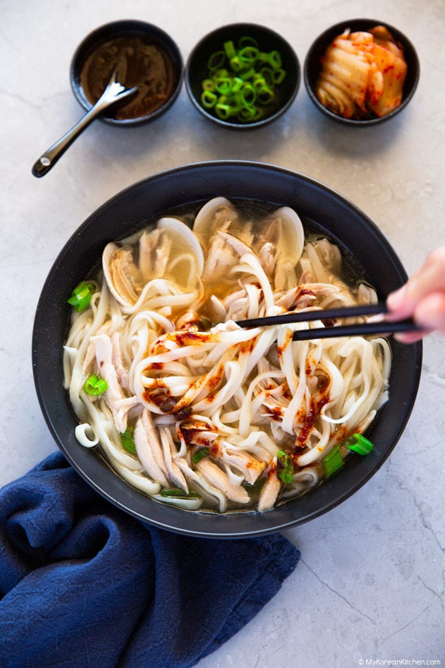 Kalguksu noodles are being held with chopsticks over a black bowl of soup, onto which red sauce is being sprinkled.