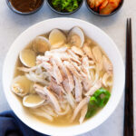 Dak kalguksu is nicely presented in a white bowl, accompanied by chopped sauce, green onions, and kimchi.