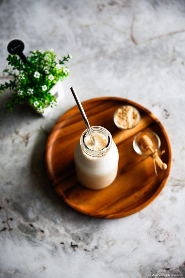 Angled shot of a misugaru latte in a glass bottle with a stainless steel straw inside. A small decorative green plant is on the left side. Also on the wooden tray are a small bowl of misugaru powder and a decorative honey spoon.