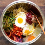 bibimbap ingredients in a large stainless steel bowl, including a sunny side egg, zucchini, spicy radish salad, bean sprout salad, and gochujang.