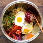 bibimbap ingredients in a large stainless steel bowl, including a sunny side egg, zucchini, spicy radish salad, bean sprout salad, and gochujang.