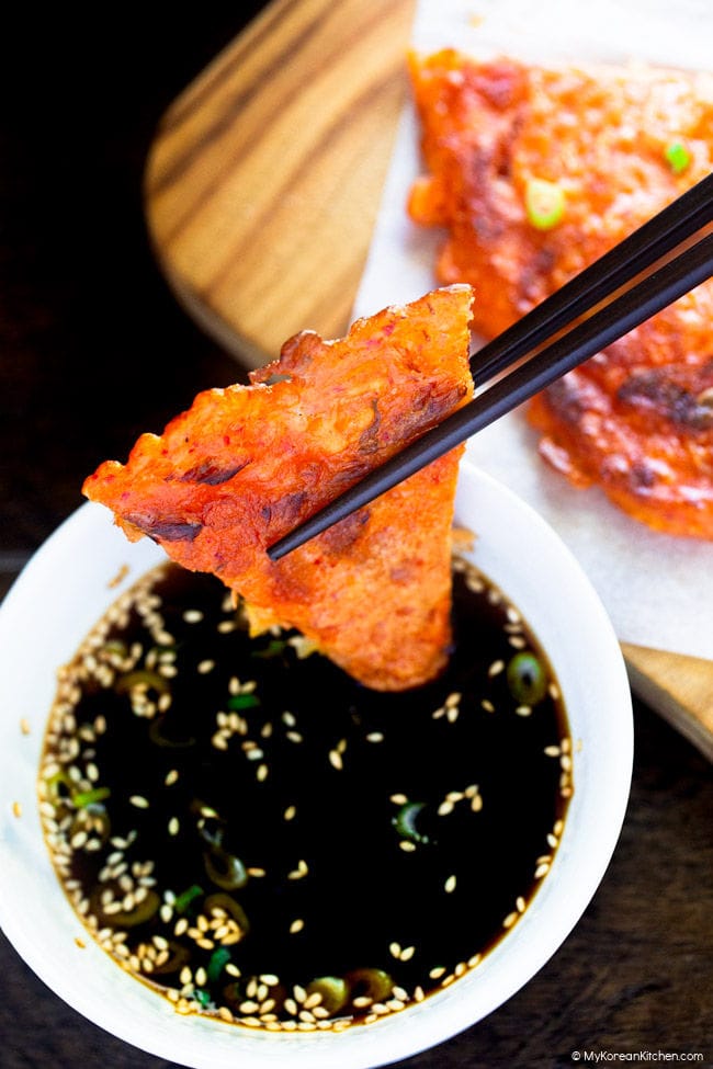 A bite of kimchi pancake held with chopsticks over dipping sauce.