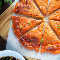 Sliced kimchi pancake into bite-sized pieces, served on a wooden board with dipping sauce and thinly sliced green onions scattered on top.