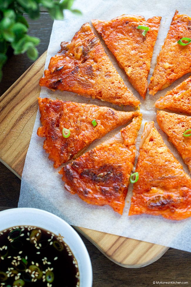 Sliced kimchi pancake into bite-sized pieces, served on a wooden board with dipping sauce and thinly sliced green onions scattered on top.