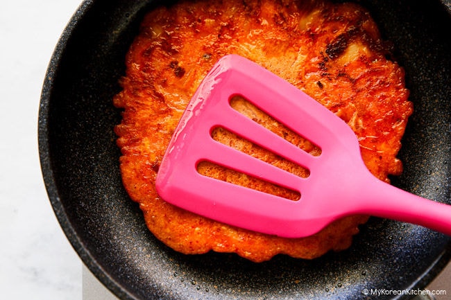 Pressing kimchi pancake in a pan with a spatula to crisp the edges.