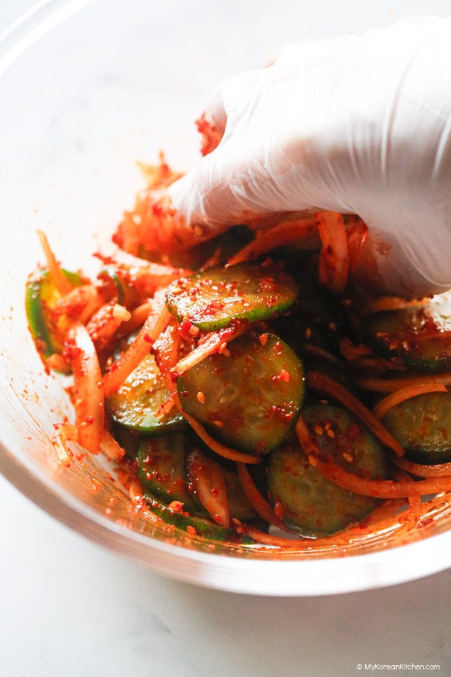 Mixing cucumber in a clear bowl with a gloved hand.
