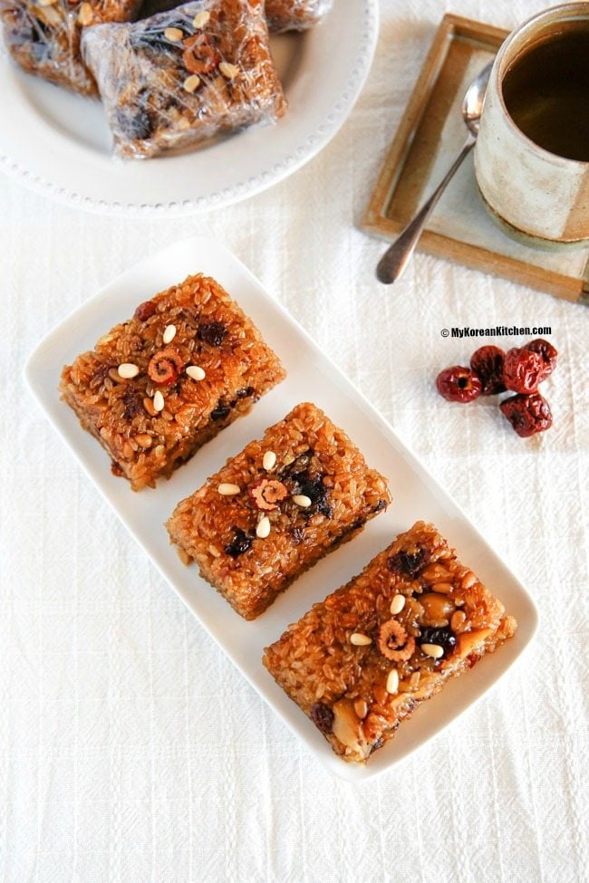 Yaksik dessert elegantly served on a rectangular white plate, with an additional three individually wrapped pieces arranged in the background on a matching round plate.