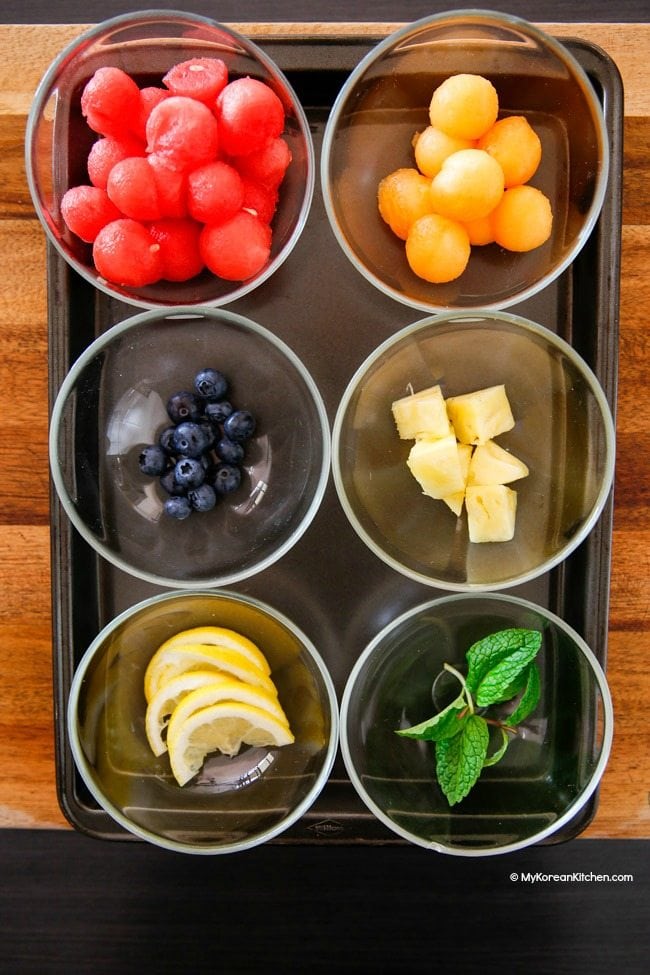 Six small glass bowls filled with fruit ingredients for Korean watermelon punch, including watermelon, lemon, pineapple, and blueberries.