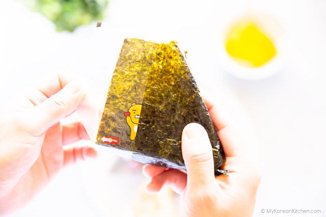 Pulling down the plastic film in the left-hand corner while holding down the triangle kimbap with right hand.