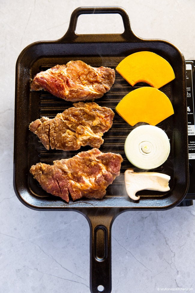 Korean BBQ Pork, sliced king oyster mushrooms, onion rings, and pumpkin on a griddle.
