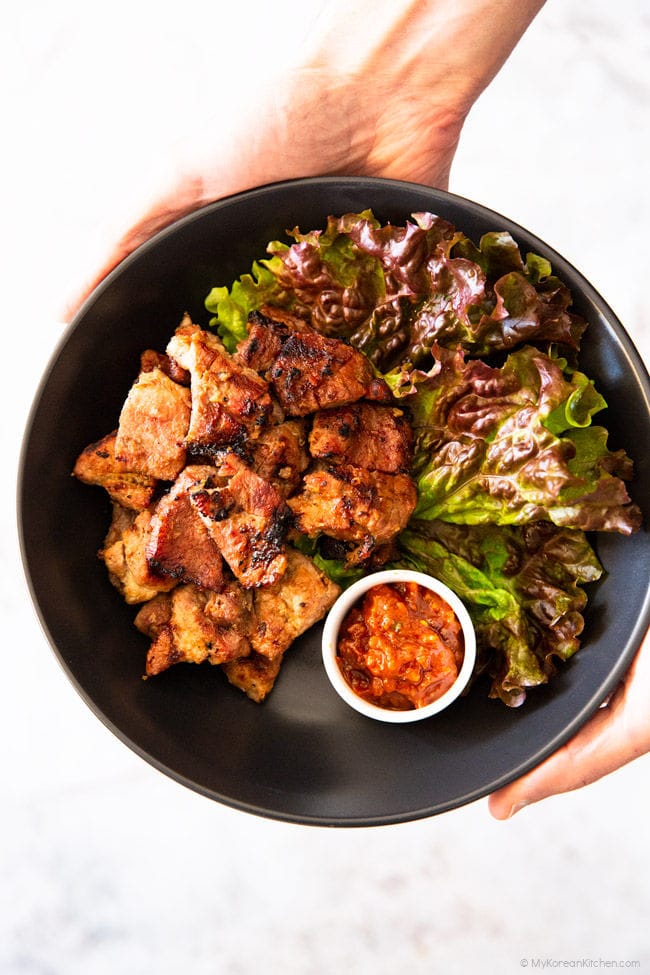 Holding a black bowl filled with Korean BBQ Pork, lettuce, and spicy dipping sauce with two hands.