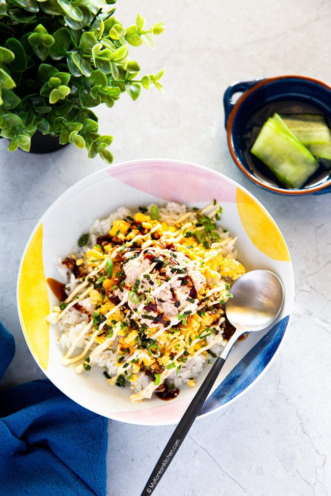 Tuna mayo rice is served in a bowl with a spoon, and pickled cucumber is served as a side dish in the corner.