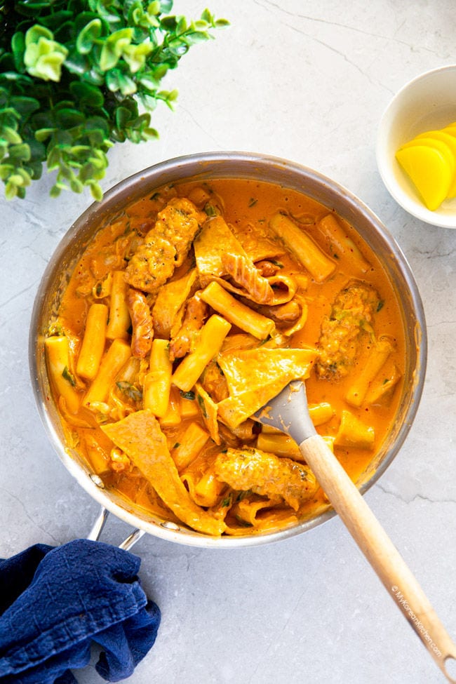 Rose tteokbokki is in a stainless skillet, with a wooden handle spatula on the side.