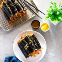 A white plate and a stainless steel tray are presented with two piles of mini kimbap, accompanied by a kimbap dipping sauce and yellow pickled radish.