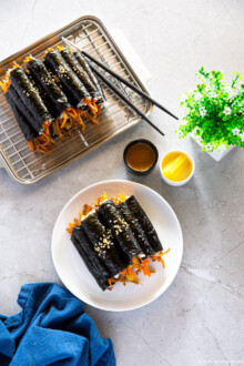 A white plate and a stainless steel tray are presented with two piles of mini kimbap, accompanied by a kimbap dipping sauce and yellow pickled radish.