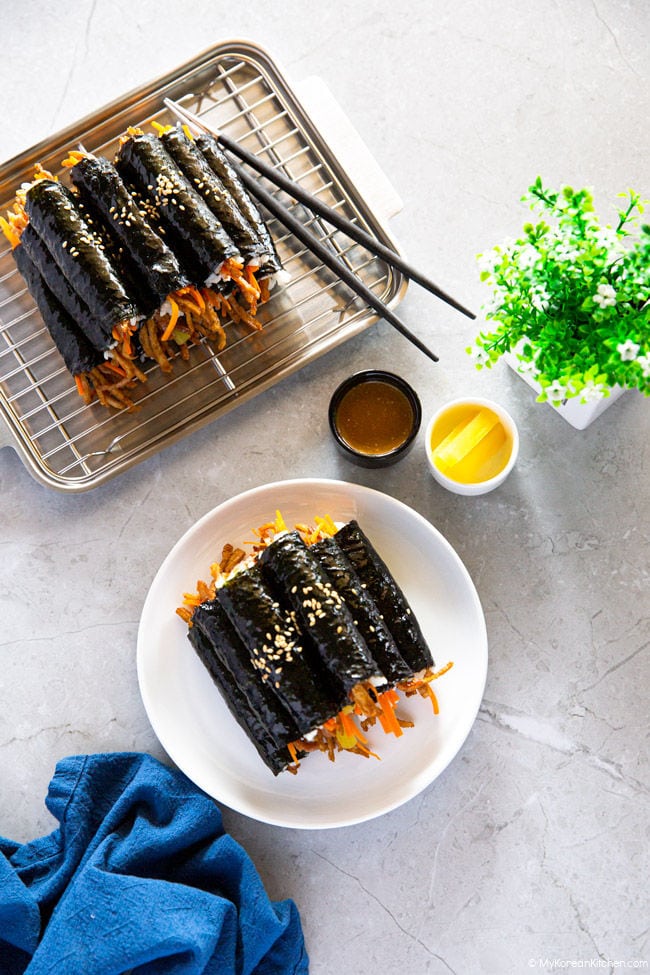 A white plate and a stainless steel tray are presented with two piles of mini kimbap, accompanied by a kimbap dipping sauce and yellow pickled radish.