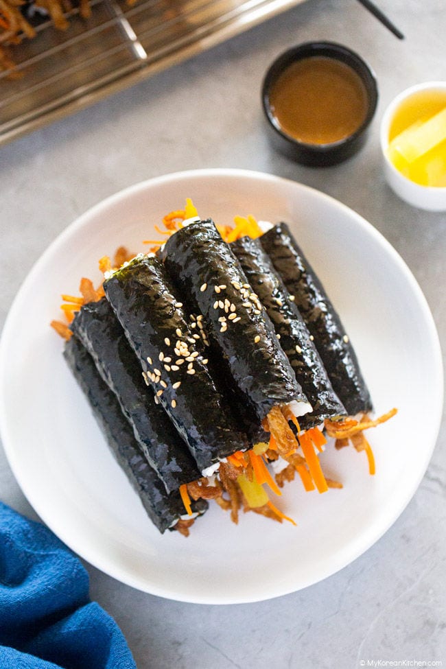 A pile of mini kimbap is served on a white plate, accompanied by a kimbap dipping sauce and yellow pickled radish.
