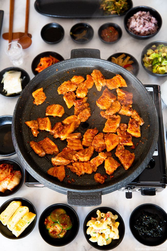 Grilled Korean spicy pork in a grill pan, surrounded by assorted Korean side dishes.