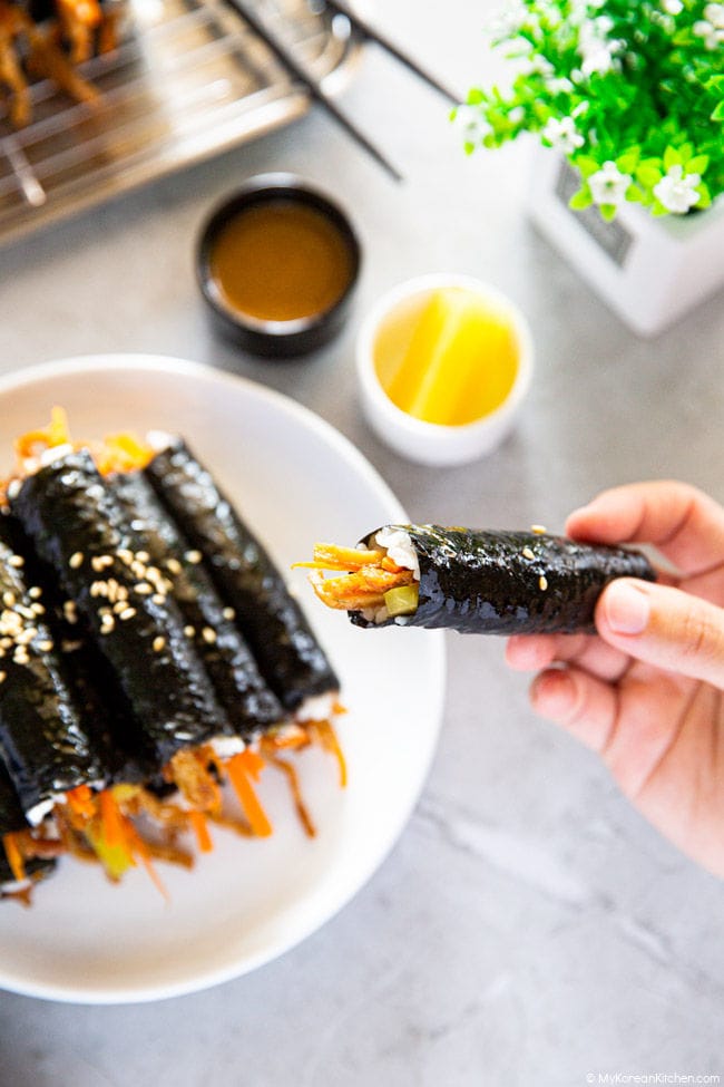 Mini kimbap in one hand, a platter of them with their accompanying kimbap sauce and yellow radish pickles in the backdrop.