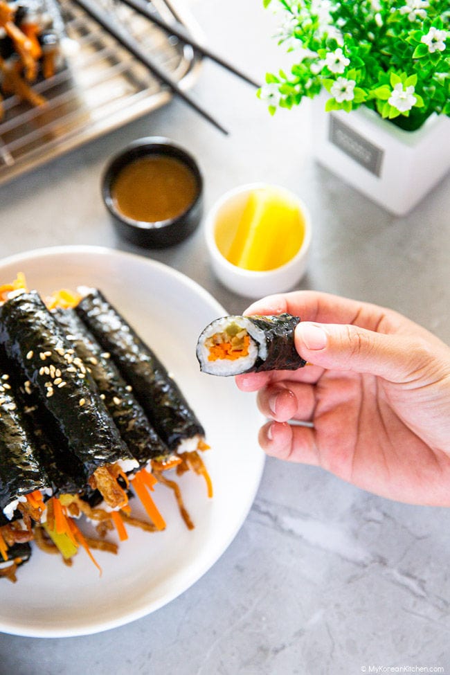 Half a mini kimbap in one hand and a platter of them with their accompanying kimbap sauce and yellow radish pickles in the backdrop.
