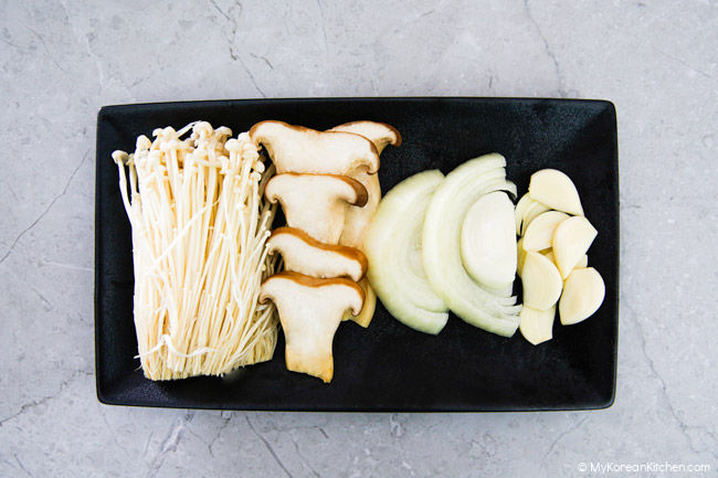 Korean BBQ vegetables are served on black rectangular plates - enoki mushrooms, king oyster mushrooms, onions, and garlic.