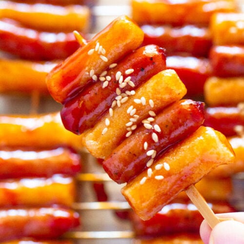 Holding one Sotteok skewer in one hand, with more Sotteok skewers visible on a plate in the background.