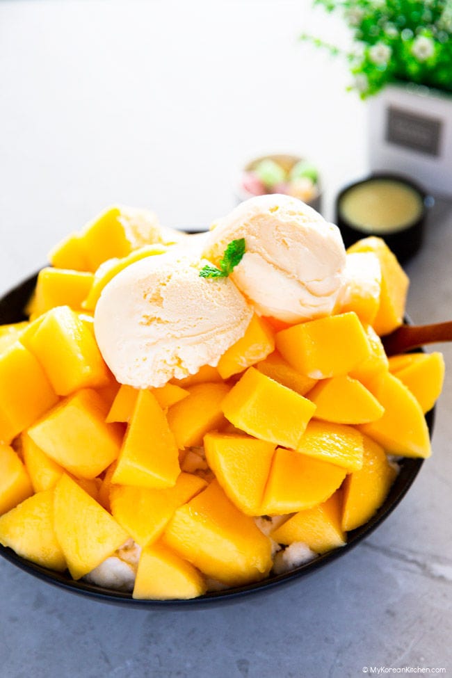 A slightly front-angled shot of a black bowl filled with a pile of sliced mangoes, topped with two scoops of ice cream.