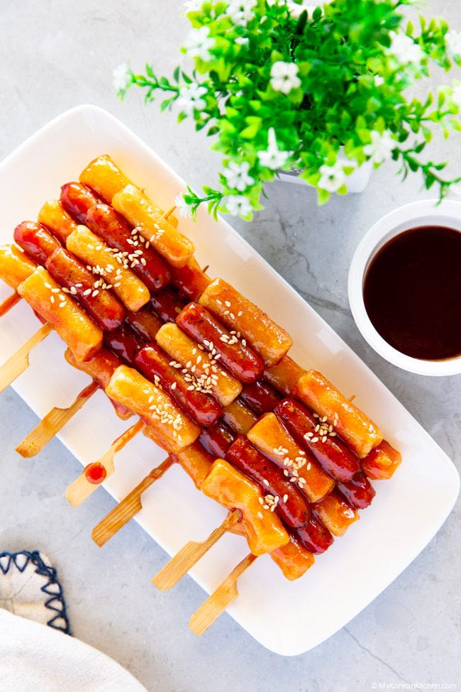 Sotteok skewers are neatly piled on a white rectangular plate. In the background, there's a pot of green plants and a bowl of extra Sotteok sauce.