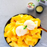 A black bowl is filled with a pile of cut mangoes, topped with two scoops of ice cream, with a text overlay saying “Mango Bingsu”.