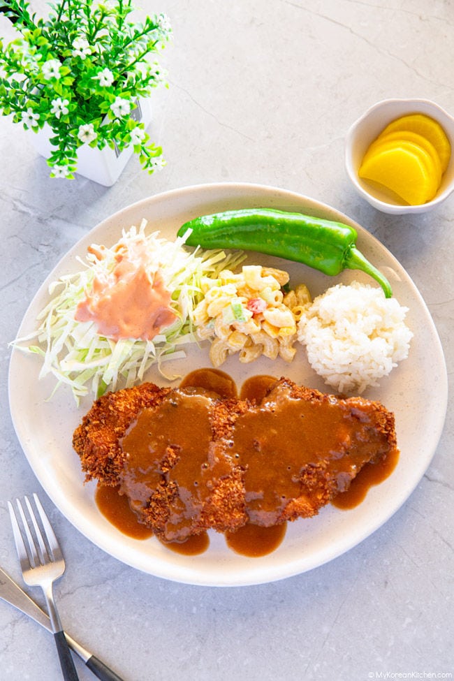 A plate filled with donkatsu covered in sauce, shredded cabbage topped with a ketchup-mayo blend, a side of green chili, macaroni salad, and a scoop of white rice.
