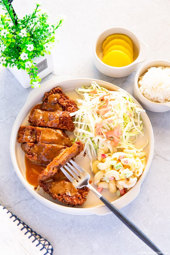 Sliced tonkatsu positioned on the left side of the plate. On the other side of the plate, there's shredded cabbage and macaroni salad. In the background, there are sliced yellow pickled radishes and rice.