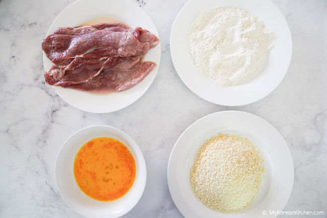 Four plates arranged on a white marble background - containing scored pork loin, flour, beaten egg, and panko breadcrumbs.