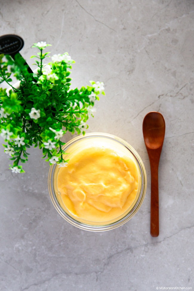 Custard in a small glass container, with a small wooden spoon on the side. In the background, there are small green plants.