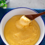 A hand holding a wooden spoon, scooping vanilla custard from a serving bowl.