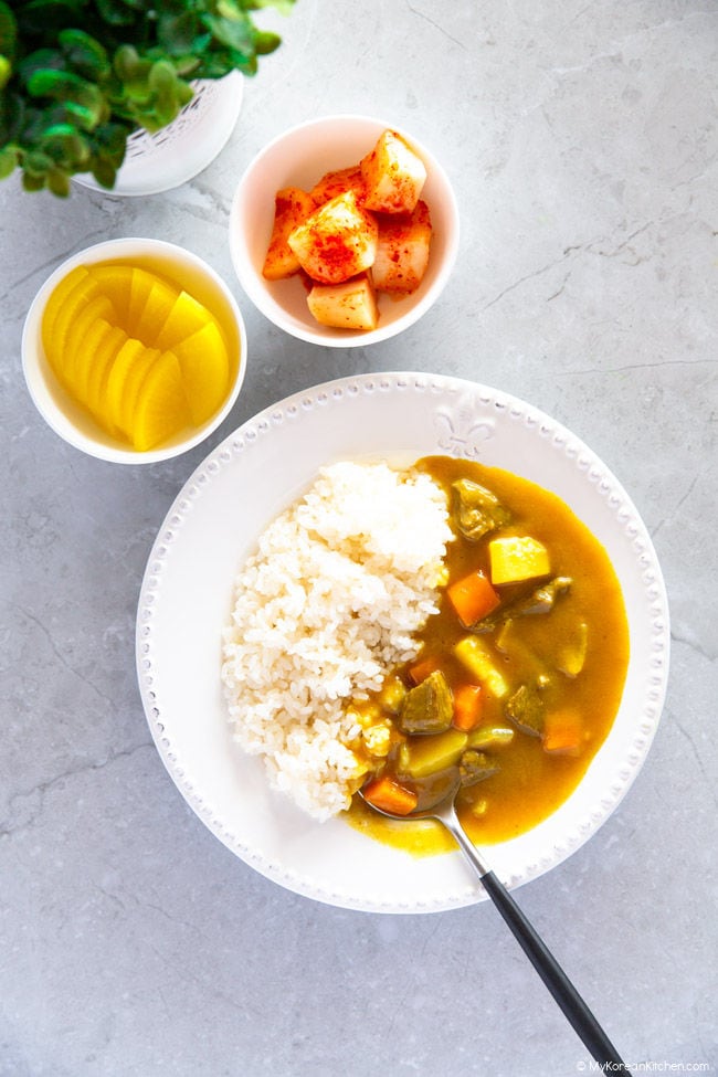 Korean beef curry is served over a bed of steamed rice on a white plate. On the side, there's yellow pickled radish and radish kimchi.