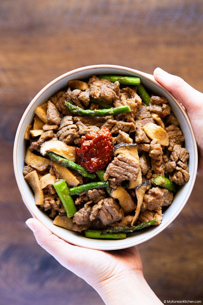 Holding a bowl of cooked Korean BBQ pork with vegetables in two hands.