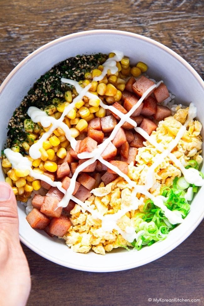 A left hand holding a bowl of Spam Egg Mayo Rice Bowl, with green onions, egg, Spam, and corn neatly arranged.