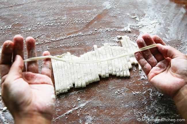 Holding a strand of knife-cut noodle by hand, with some sliced noodles on a floured cutting board in the background